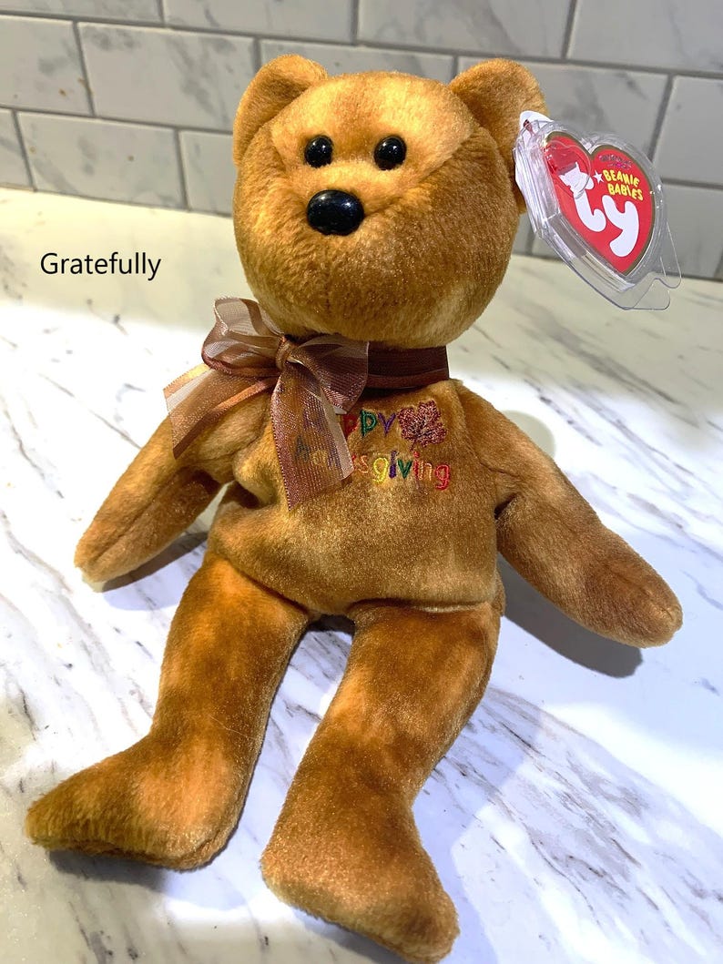 May include: A brown Ty Beanie Baby bear plush toy with a brown ribbon and the word "Thanksgiving" embroidered on its chest. The bear has a black nose and a red heart-shaped tag.