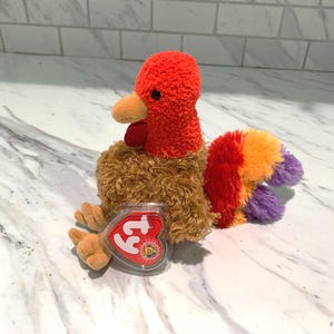 May include: A plush turkey toy with a red head and wattle, orange beak, and brown body. The tail feathers are a mix of red, orange, and purple. A heart-shaped tag is attached to the toy.
