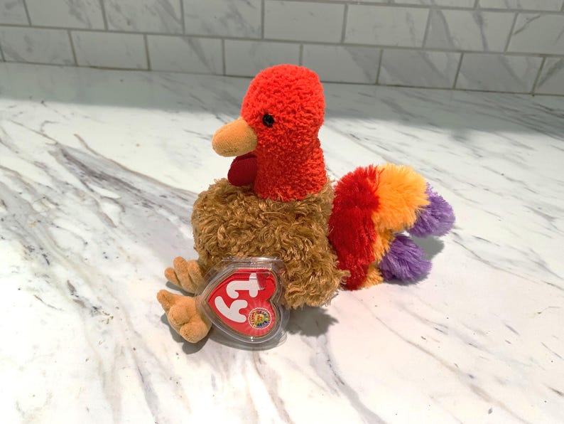 May include: A plush turkey toy with a red head and wattle, orange beak, and brown body. The tail feathers are a mix of red, orange, and purple. A heart-shaped tag is attached to the toy.