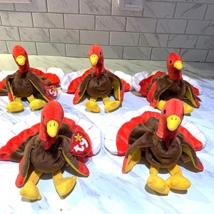 May include: Five plush turkey toys with red, brown, and yellow details. Each turkey has a red head, brown body, and red and white tail feathers. The turkeys are sitting on a white and gray surface.