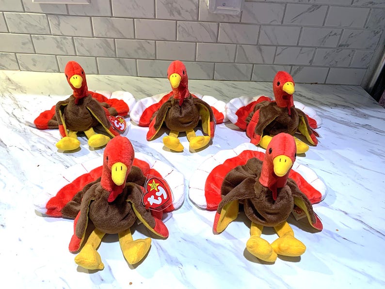 May include: Five plush turkey toys with red, brown, and yellow details. Each turkey has a red head, brown body, and red and white tail feathers. The turkeys are sitting on a white and gray surface.