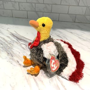 May include: A plush turkey toy with a yellow head, orange beak and feet, and a red wattle. The body is a mix of gray, white, and red plush. A red heart-shaped tag is attached to the toy.