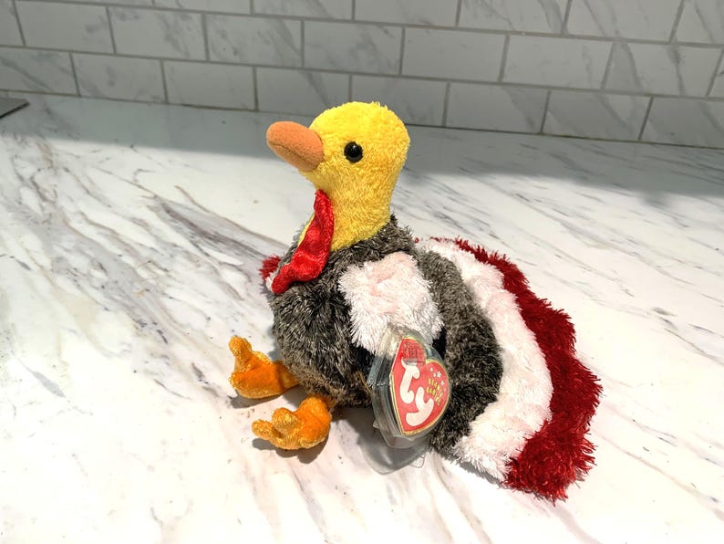 May include: A plush turkey toy with a yellow head, orange beak and feet, and a red wattle. The body is a mix of gray, white, and red plush. A red heart-shaped tag is attached to the toy.