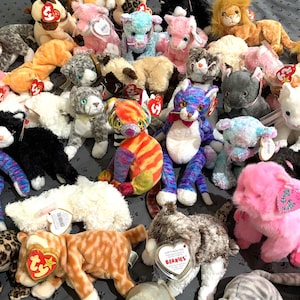 May include: A collection of colorful plush toys, mostly cats, with red and white Ty tags. The toys are arranged on a gray and white patterned surface.