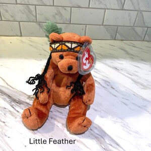 May include: A plush brown bear toy named "Little Feather" with a Native American-style headband and feather. The bear has black yarn hair and a red heart-shaped tag.