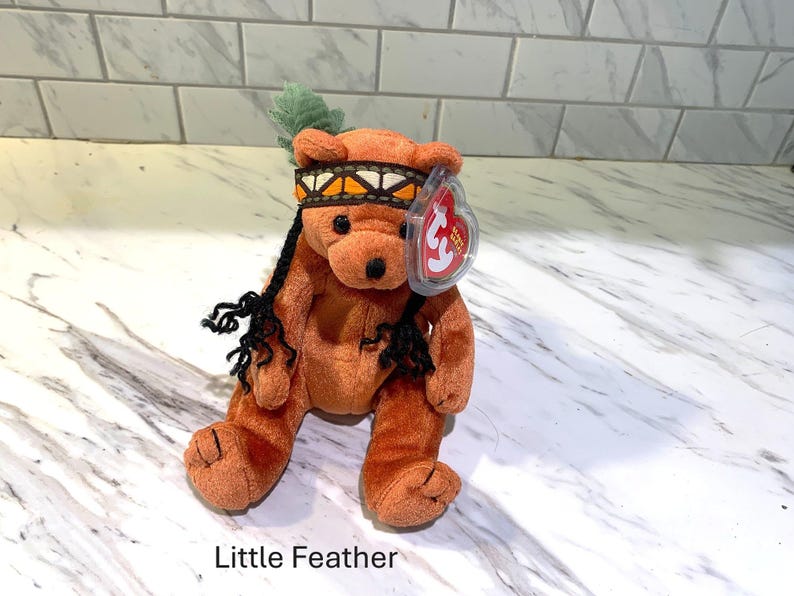 May include: A plush brown bear toy named "Little Feather" with a Native American-style headband and feather. The bear has black yarn hair and a red heart-shaped tag.
