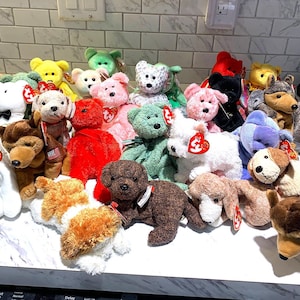 May include: A collection of plush stuffed animals, including bears and dogs, in various colors such as white, pink, brown, and green. Each animal has a tag attached. The toys are arranged on a white surface.