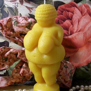以下が含まれることがあります： テクスチャ加工された頭を持つ女性の形をした黄色の蜜蝋のろうそく。ろうそくは、真珠のネックレスと花柄の布のある白い表面の上に置かれています。