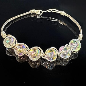 May include: A silver wire bangle bracelet with iridescent glass beads. The bracelet is made of silver wire and has a simple design. The beads are iridescent and have a rainbow effect.