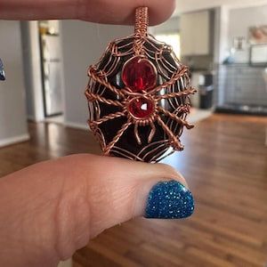 Puede incluir: Un colgante hecho a mano con una piedra oscura envuelta en alambre de cobre, con un diseño de araña y telaraña. La araña tiene dos acentos de piedras preciosas rojas. El colgante se sujeta con un lazo de alambre de cobre.