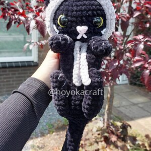 May include: A black crochet cat plushie with a grey and red horned hood and a white scarf. The cat has large yellow eyes and a white nose. The text "@hoyokacraft" is visible on the plushie.
