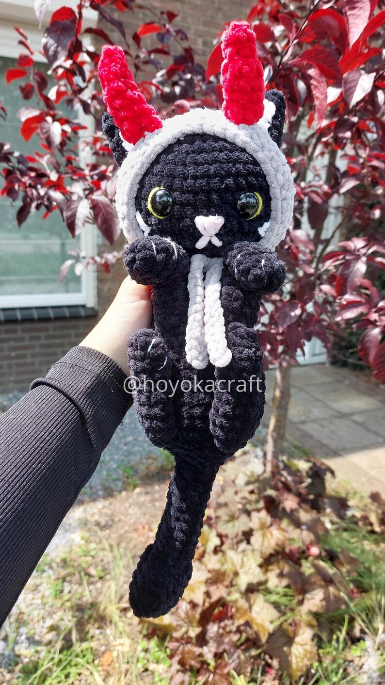 May include: A black crochet cat plushie with a grey and red horned hood and a white scarf. The cat has large yellow eyes and a white nose. The text "@hoyokacraft" is visible on the plushie.