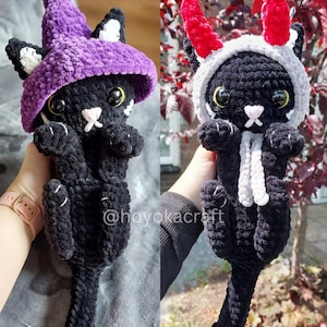 Peut inclure: Deux peluches de chat en crochet noir faites à la main. L'un porte un chapeau de sorcière violet avec des accents blancs, l'autre porte un sweat à capuche gris avec des cornes rouges. Les deux ont des marques blanches et de grands yeux ronds. Les peluches sont tenues sur un fond flou.