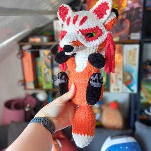 May include: A plush fox toy crafted from orange, white, and black yarn. The fox features a white mask with red markings over its face, black paws, and a long tail with a white tip. The toy has a soft, textured appearance with red tassels.