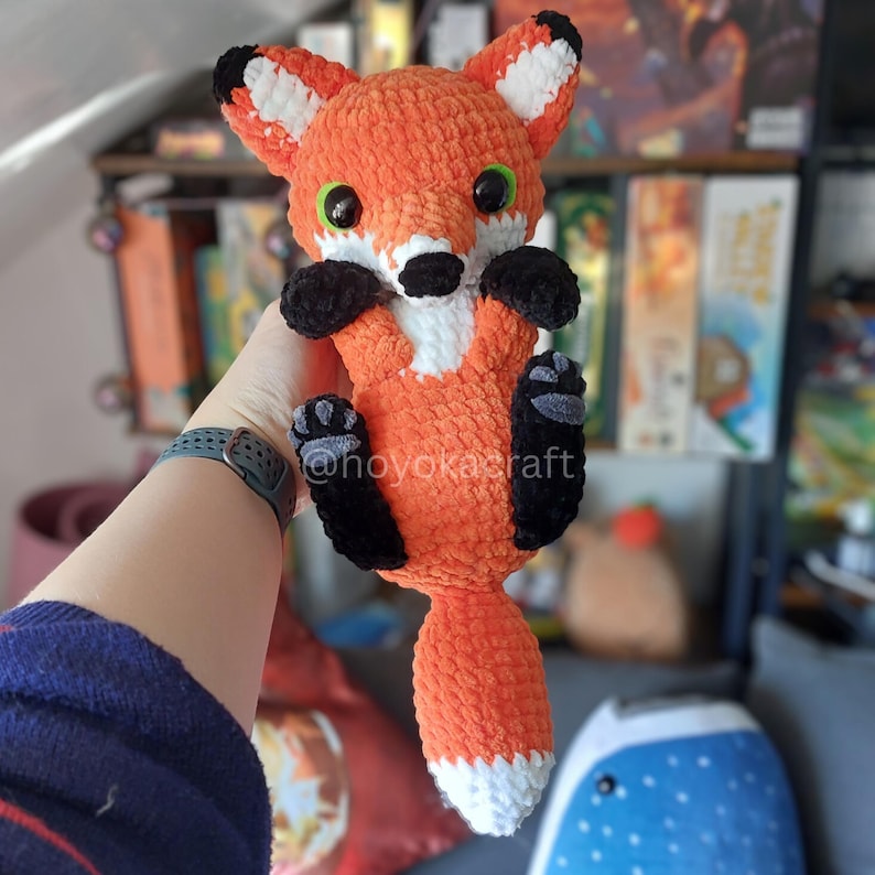 May include: A crocheted fox plush toy featuring bright orange yarn, white accents on the ears, chest, and tail tip, and black yarn for the paws and nose. The fox has large, round, green plastic eyes and a soft, textured appearance throughout.