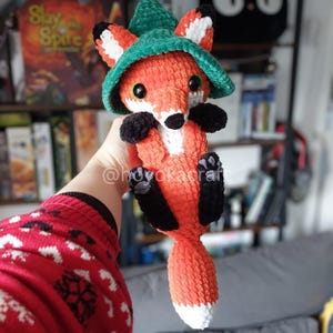 May include: A handmade orange and white crochet fox wearing a green hat. The fox has black paws and a white chest. The fox is holding its paws up. The fox has large, round eyes.