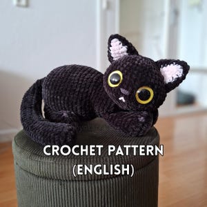 Könnte beinhalten: Ein gehäkeltes schwarzes Katzen-Stofftier mit großen gelben Augen, einer kleinen grauen Nase und hellrosa Innenohren. Die Katze liegt mit eingerolltem Schwanz. Der Text CROCHET PATTERN (ENGLISH) ist in weißen Buchstaben auf dem Bild zu sehen.