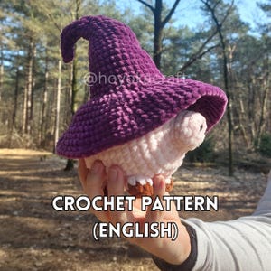 Puede incluir: Una figura de hongo de ganchillo con un sombrero morado y un cuerpo blanco y rosa. El texto "CROCHET PATTERN (ENGLISH)" se muestra en la parte inferior de la imagen. La figura se sostiene en una mano.