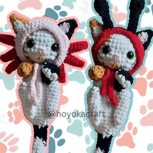 Può includere: Due peluche di gatto fatti a mano all'uncinetto, uno rosa e l'altro rosso, ciascuno con cappuccio e branchie simili a quelle di un axolotl. I gatti hanno corpi bianchi, dettagli neri e arancioni e grandi occhi verdi. Lo sfondo presenta impronte di zampe.