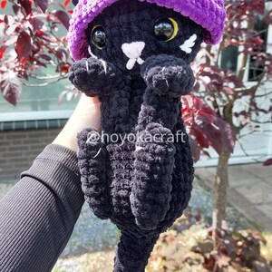 May include: A handmade black crochet cat doll wearing a purple witch hat with white accents. The cat has large, round eyes and a white nose and whiskers. The doll is held up by a person's hand.