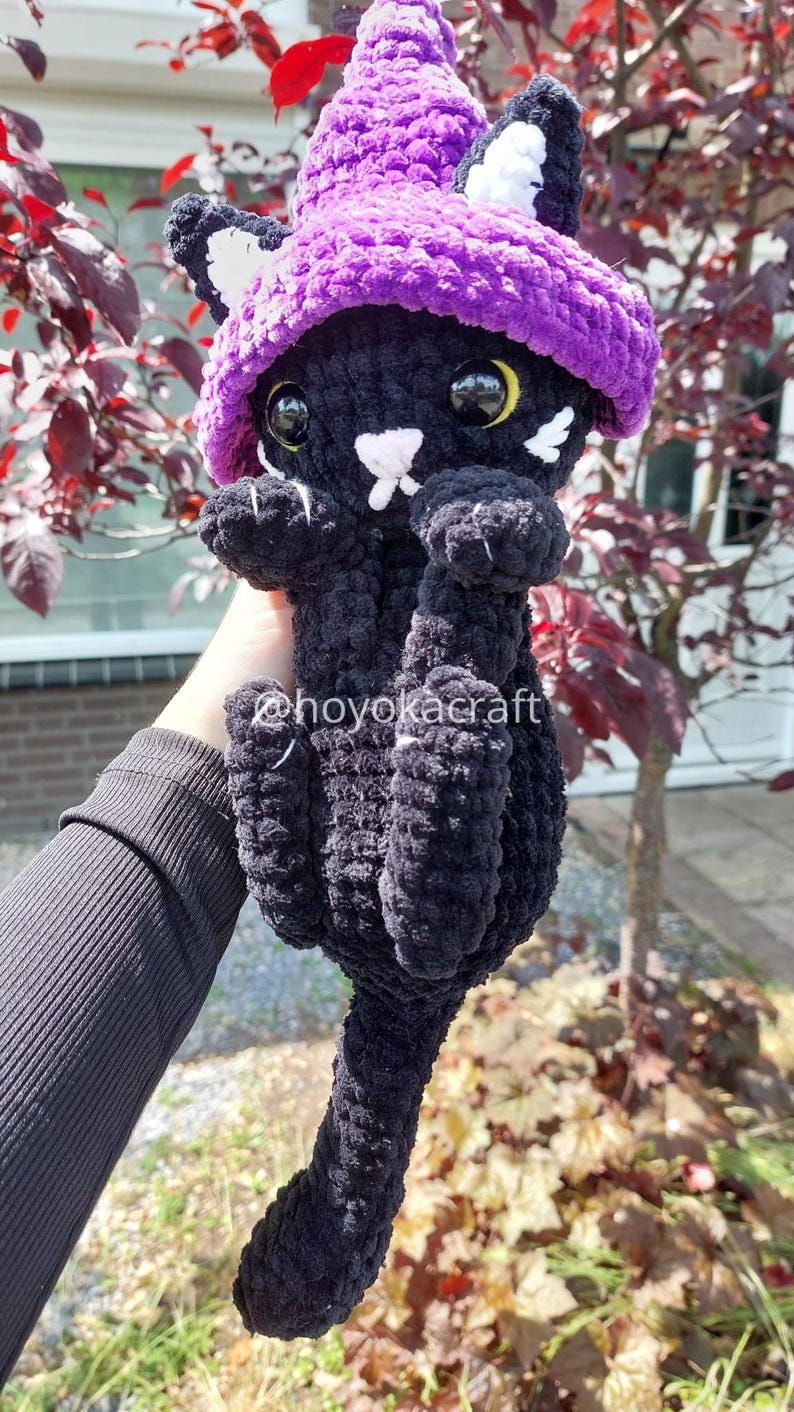 May include: A handmade black crochet cat doll wearing a purple witch hat with white accents. The cat has large, round eyes and a white nose and whiskers. The doll is held up by a person's hand.