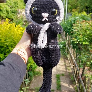 May include: A handmade black crochet cat doll wearing a white and grey hood with red horns. The cat has yellow eyes and a white nose. The doll has a white scarf and the text "@hoyokacraft".