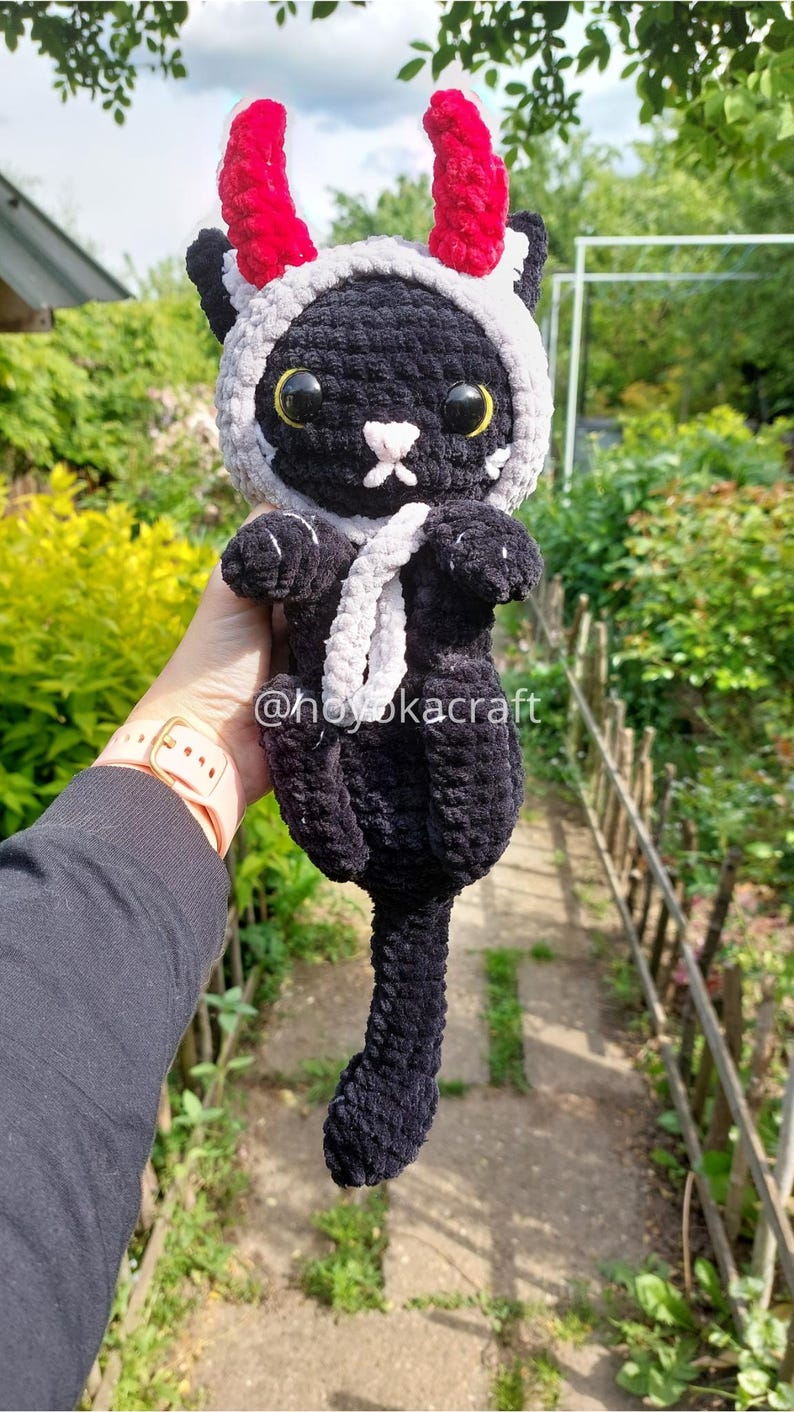 May include: A handmade black crochet cat doll wearing a white and grey hood with red horns. The cat has yellow eyes and a white nose. The doll has a white scarf and the text "@hoyokacraft".