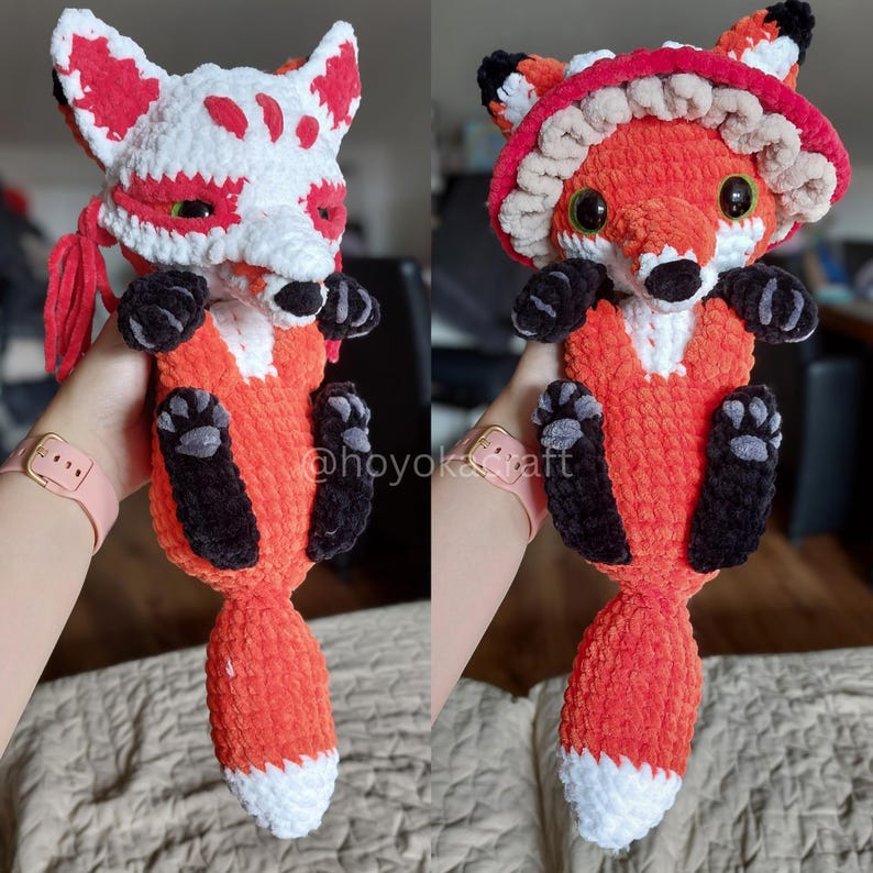 May include: A plush crochet fox toy featuring a soft orange and white body with black paws and a white-tipped tail. The fox is shown in two configurations: one wearing a white fox-themed mask and another wearing a red and beige mushroom-style hat.