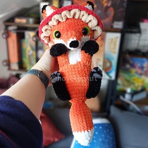 May include: A plush crochet fox toy featuring bright orange fur, a white chest patch, and black paws. The fox wears a red and beige mushroom-shaped hat with a ruffled edge. It has large green eyes, a black nose, and a long tail with a white tip.