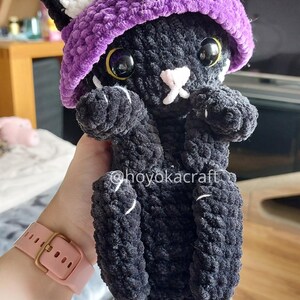 May include: A black crochet cat wearing a purple witch's hat. The cat has white details on its ears and paws, with yellow eyes and a pink nose. The doll is soft and plush, suitable as a child's toy or a unique home decoration.