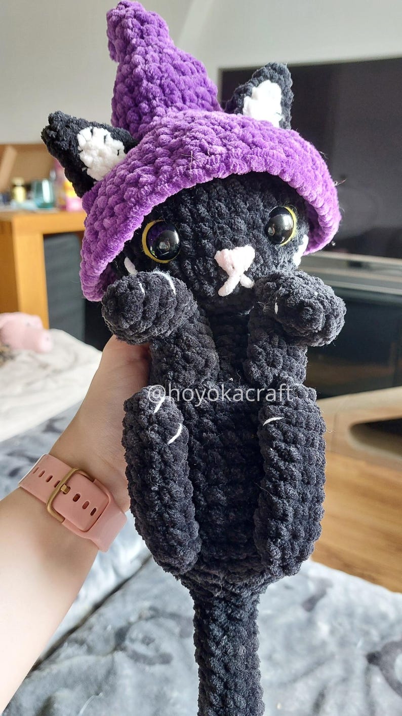 May include: A black crochet cat wearing a purple witch's hat. The cat has white details on its ears and paws, with yellow eyes and a pink nose. The doll is soft and plush, suitable as a child's toy or a unique home decoration.