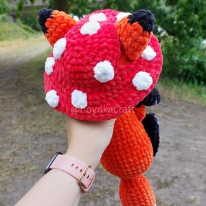 May include: A plush crochet hat shaped like a red mushroom cap with white spots and two orange fox ears with black tips. The hat features a soft, textured yarn construction and includes the text @noyokacraft printed on the surface of the fabric.