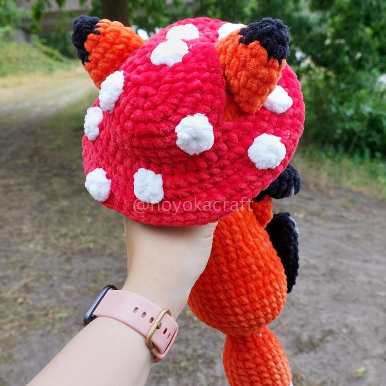 May include: A plush crochet hat shaped like a red mushroom cap with white spots and two orange fox ears with black tips. The hat features a soft, textured yarn construction and includes the text @noyokacraft printed on the surface of the fabric.