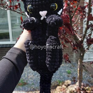 May include: A handmade black crochet cat with white detailing on the ears and paws. The cat has large yellow eyes and a pink nose. The text "@hoyokacraft" is visible. A plush toy cat.