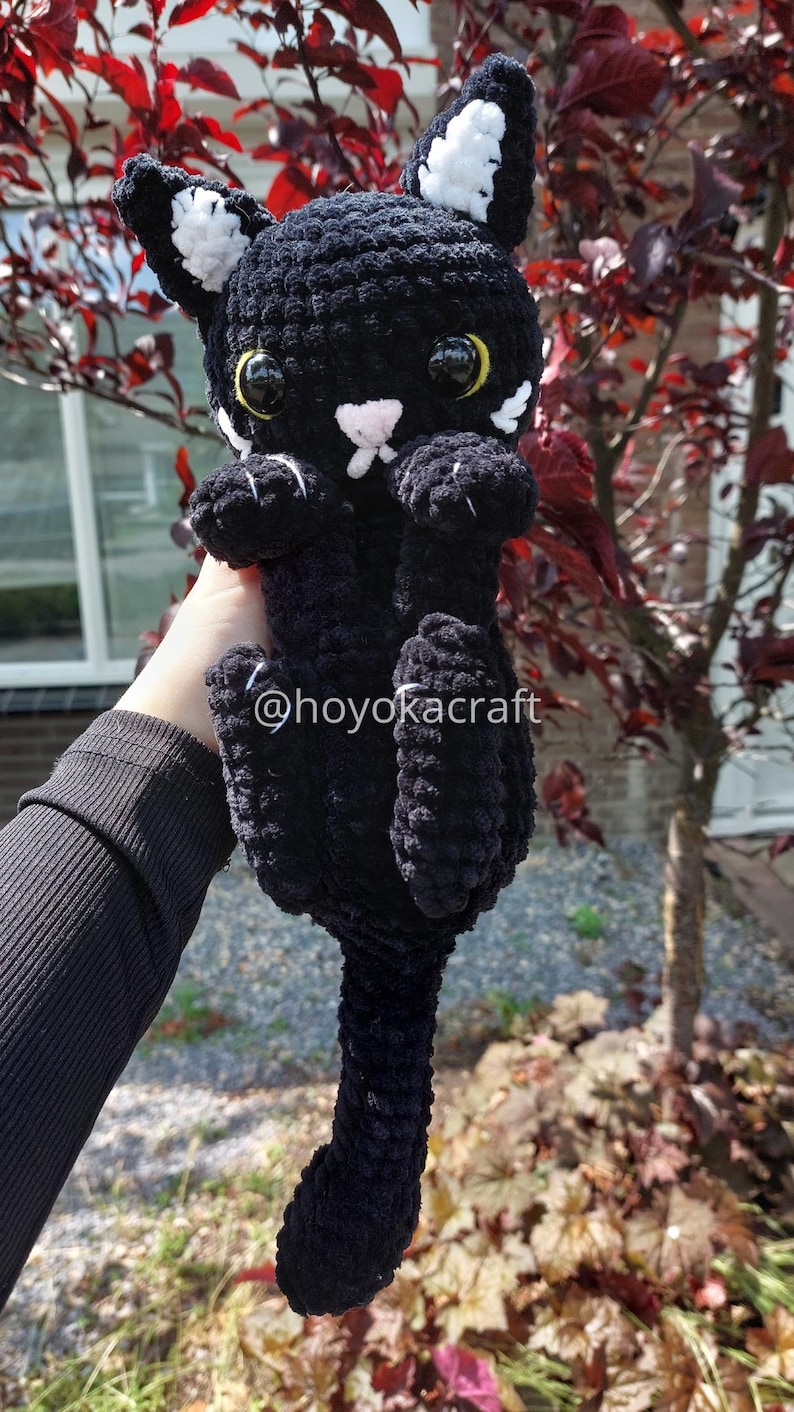 May include: A handmade black crochet cat with white detailing on the ears and paws. The cat has large yellow eyes and a pink nose. The text "@hoyokacraft" is visible. A plush toy cat.