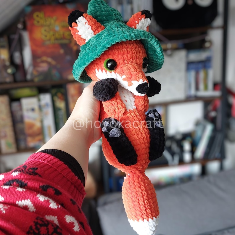 May include: A handmade orange fox plushie with a green hat, black paws, and white accents. The fox has large black eyes and is holding a pose. The plushie is made of soft, textured yarn.