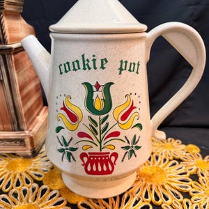 May include: Cream-colored ceramic cookie pot with lid. The pot features a floral design in red, yellow, and green, with the words "cookie pot" in green lettering. The pot has a handle and spout.