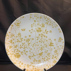 May include: White ceramic plate with a yellow floral design featuring flowers, leaves, and butterflies. The plate is round and has a slightly raised edge. The design is a repeating pattern across the plate's surface.