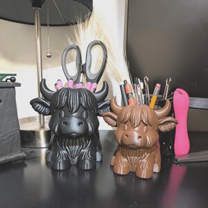 May include: Two Highland cow-shaped desk organizers, one black and one brown, sit side-by-side. The black cow holds scissors and pink pens, while the brown cow contains pencils and pens. Both have horns and shaggy fur details, adding a whimsical touch.