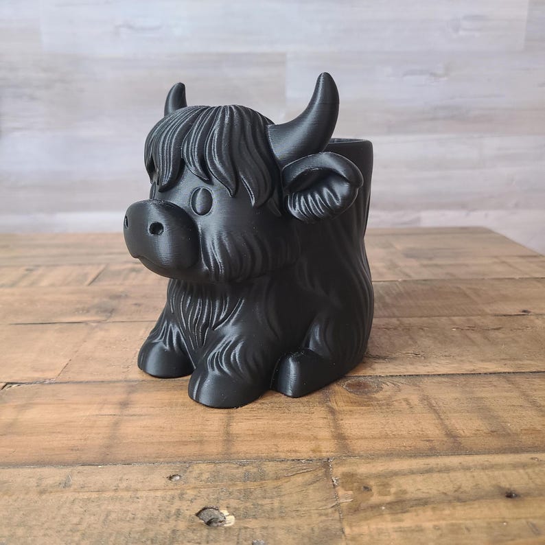 May include: A black Highland cow planter, featuring a detailed design with horns, shaggy fur, and a friendly face. The planter is made of a solid material and sits on a wooden surface. The cow is in a sitting position.