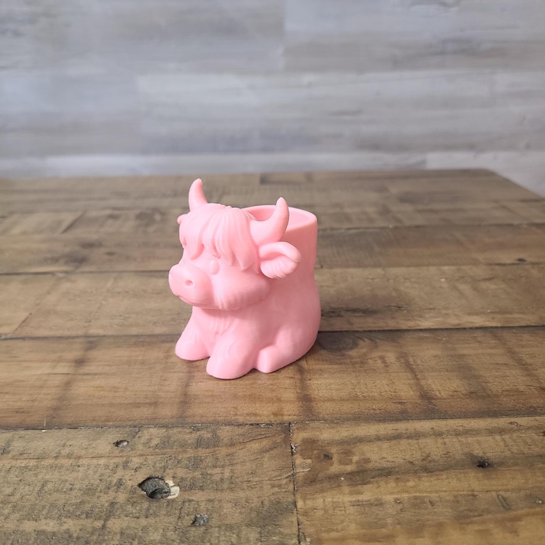 May include: A pink Highland cow planter made of ceramic. The planter is shaped like a cow with horns, a fluffy fringe, and a round body. The planter is sitting on a wooden surface.