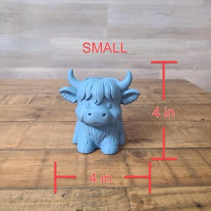 May include: A small, light blue Highland cow figurine. The figurine is 4 inches tall and 4 inches wide. The word "SMALL" is written above the figurine. The cow has horns, shaggy fur, and a friendly expression.