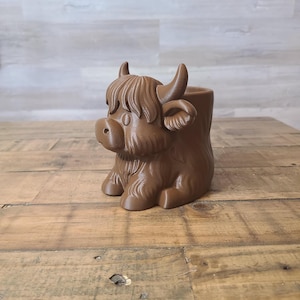 May include: A brown Highland cow planter with a detailed design. The planter features a sitting cow with horns, a fluffy fringe, and a round body. The planter is made of a solid material and is suitable for small plants or succulents.
