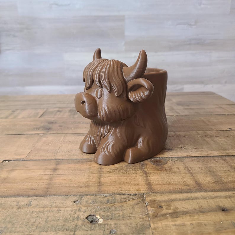 May include: A brown Highland cow planter with a detailed design. The planter features a sitting cow with horns, a fluffy fringe, and a round body. The planter is made of a solid material and is suitable for small plants or succulents.