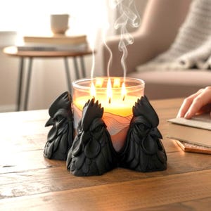 May include: A lit candle in a clear glass jar with three wicks, surrounded by three black rooster figurines. The candle is on a wooden table, with smoke rising from the flames. The candle has a yellow hue.