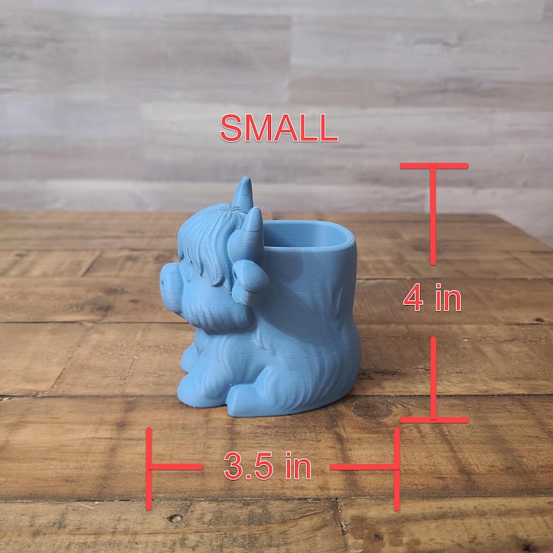 May include: A light blue, 3D-printed planter shaped like a Highland cow. The planter is 4 inches tall and 3.5 inches wide. The word "SMALL" is printed in red on the top. The planter is sitting on a wooden surface.