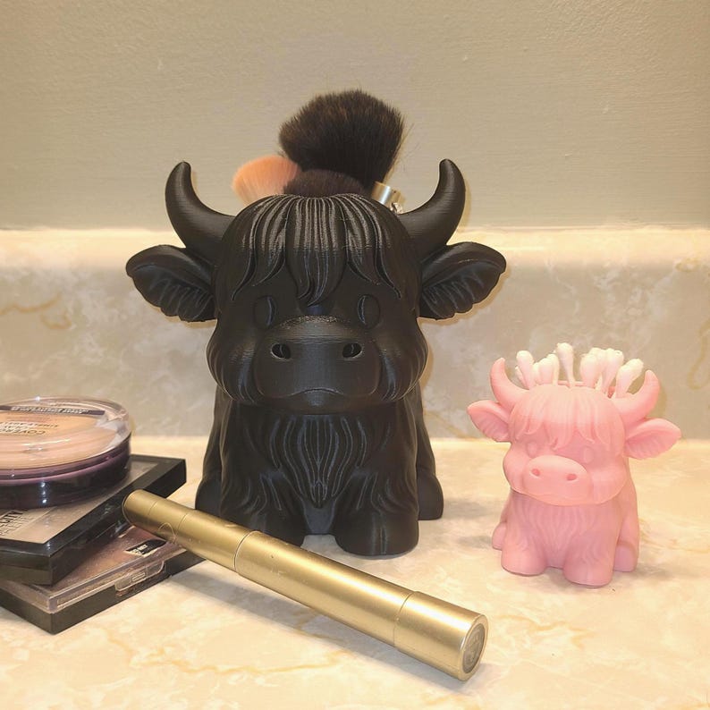 May include: A black Highland cow-shaped makeup brush holder with a dark brown brush and several smaller brushes. A pink Highland cow figurine is next to the holder. A gold-colored mascara tube and makeup compacts are in the foreground.