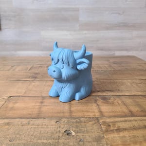 May include: A light blue Highland cow planter, featuring a detailed sculpted design. The planter has a small opening at the top for plants and is sitting on a wooden surface. The cow has horns and a fluffy fringe.