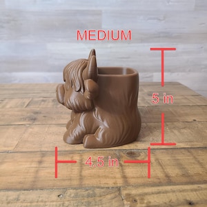 May include: A brown, medium-sized planter shaped like a Highland cow. The planter is 5 inches tall and 4.5 inches wide. The word "MEDIUM" is printed in red. The planter is made of a solid material and has a textured appearance.