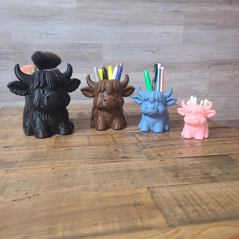 May include: Four Highland cow-shaped desk organizers in black, brown, blue, and pink. Each has a pen holder design, with pens and a makeup brush in some. The organizers are made of a solid material and sit on a wooden surface.